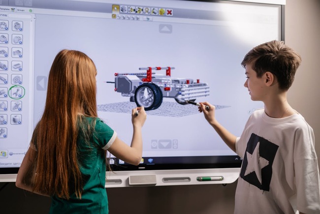 Doi adolescenți care arată cu markerul spre un proiect de robotică