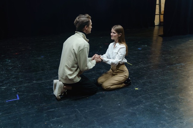 Doi actori pe scenă care stau în genunchi și se țin de mână